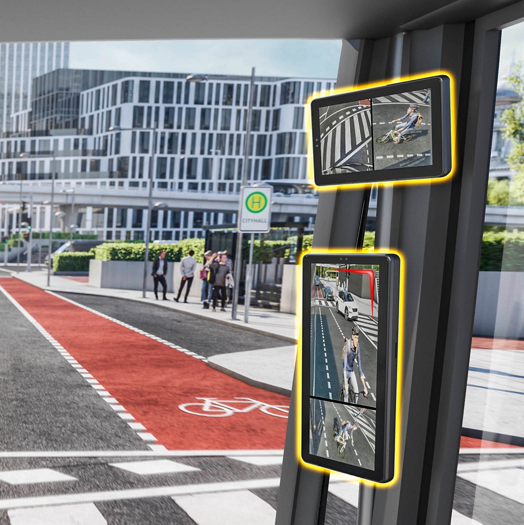 The view of a monitor mounted on the A column of a commercial vehicle. The monitor shows the scene right next to the vehicle, usually the blind spot. The image shows a cyclist from two different angles. To the front, there is a red bike line and a bus stop.